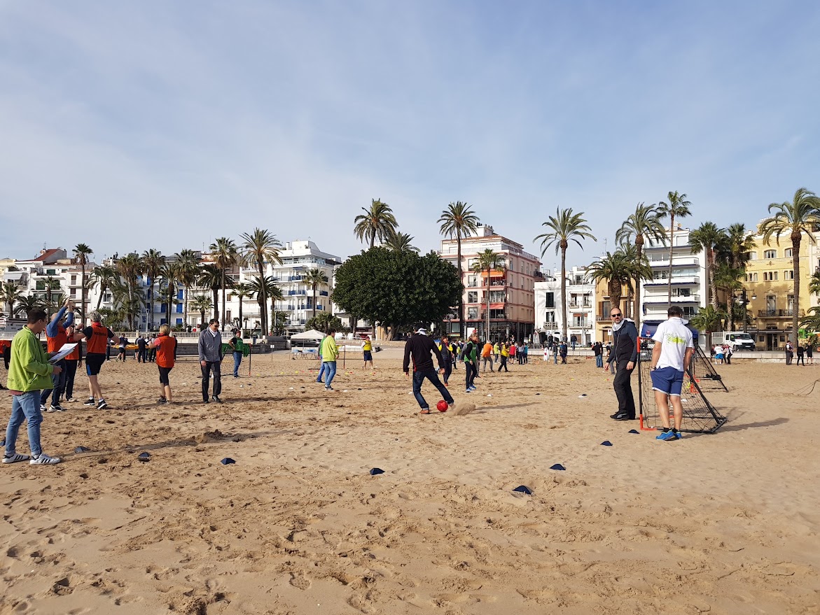 Group participating in a team-building activity on a sunny beach in Barcelona, surrounded by palm trees and vibrant coastal architecture.